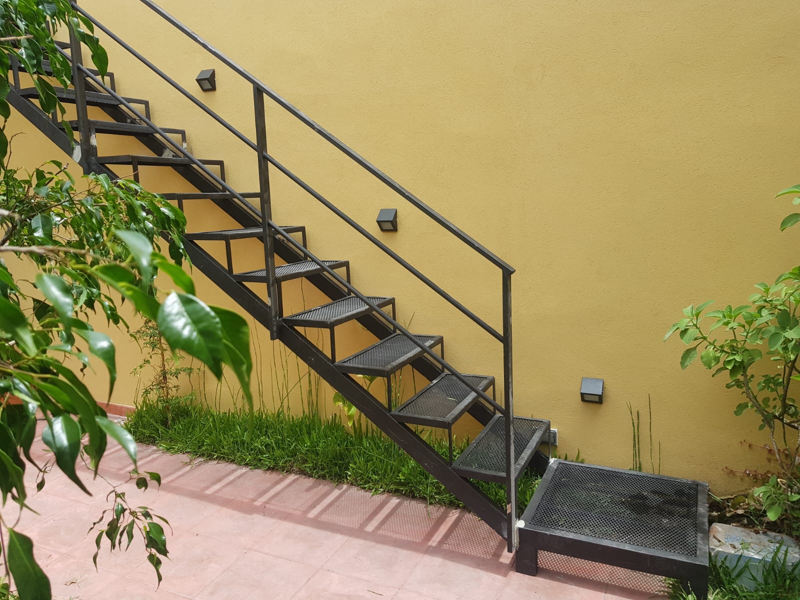 Escalera recta negra exterior junto a pared amarilla y jardín