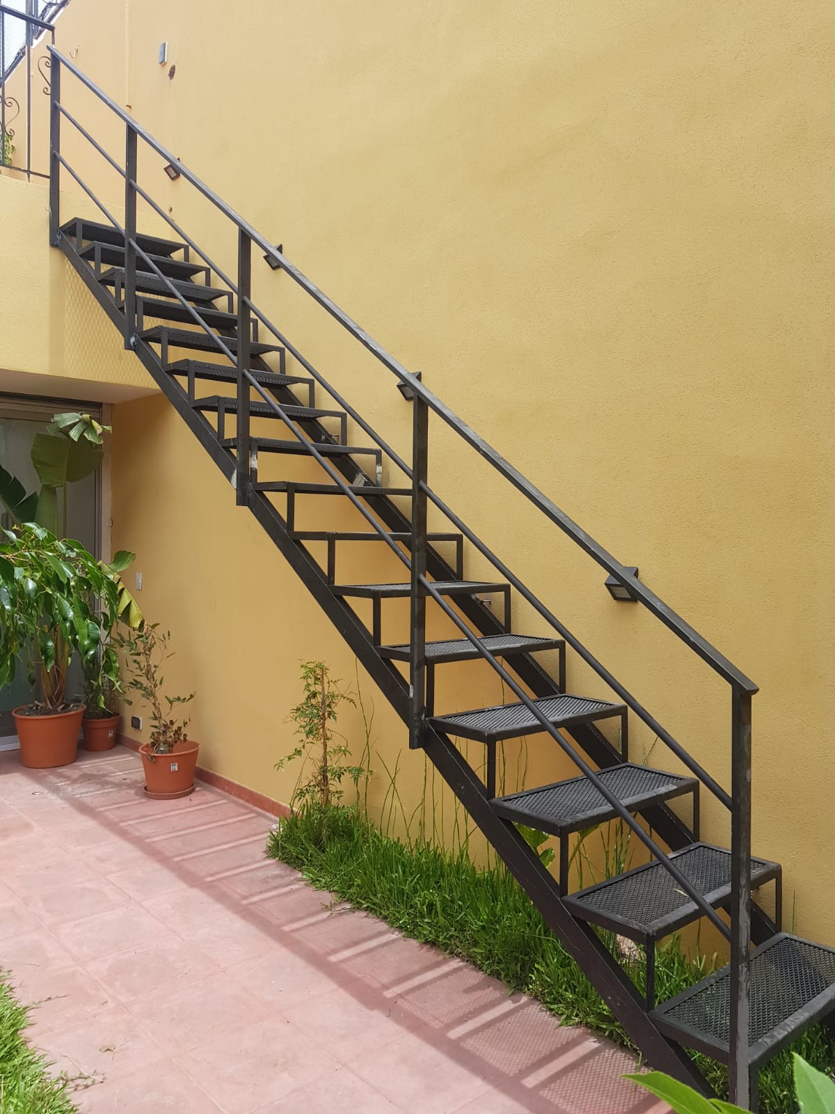 Escalera recta negra exterior junto a pared amarilla y jardín